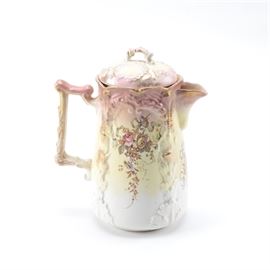 Chinese E&M Porcelain Pitcher: A Chinese E&M porcelain pitcher. This pitcher features a cylindrical body with and embossed foliate design and a squared handle. This pitcher is glazed in a pink to cream-colored glaze and features a floral design with gilt accents and trim. The base of this piece is marked “E&M”.