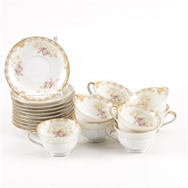Noritake Teacups and Saucers: A selection of Noritake teacups and saucers. This selection includes nine teacups and ten saucers with a floral and scroll design and gilt trim. Each item is marked “Noritake Japan” on the base.
