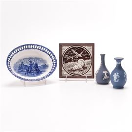 Selection of Wedgwood Featuring Jasperware: A selection of Wedgwood. This group includes two Jasperware bud vases with white molded neoclassical style figures of cupid with birds and leaf garlands and an oval earthenware serving bowl with a basket weave design, pierced edges, and a blue depiction of a ship in a harbor to the center. These pieces are marked “Wedgwood” to the underside. The lot also includes a framed square tile with a depiction of a dead deer and flying eagle. It reads “Where the Carcase is, There will the Eagles be Gathered Together”. It is presented in a wooden frame.