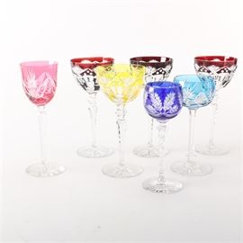 Colored Cut Glass Stemware: A collection of assorted cut colored glass stemware. This selection features a blue glass with a clear cut leaf motif on the sides; a light blue glass with a fan pattern; a yellow glass and a pink glass with various fan motifs; and three matching cranberry red glasses. All seven glasses feature clear glass stems with starburst patterns on the underside of the base.