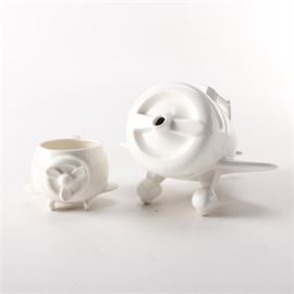 Airplane Teapot with Mug From L. Stuart Taylor and Vandor Imports: An airplane teapot and mug designed by L. Stuart Taylor, produced by Vandor Imports. This set includes a mug with small feet shaped like airplane wheels, and a front adorned with a propeller design. The teapot is shaped like an airplane, with the spout protruding from the center of the propeller, and the lid where the cockpit would be. There are markings and decals to the underside of the pieces, indicating they were produced by Vandor Imports in 1978, and designed by L. Stuart Taylor.