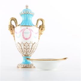 Porcelain Décor: A pair of porcelain decorative items. This pair of decorative porcelain items features an ash receiver with a gold tone rim decorated with a blue floral design to the center marked “Limoges France”. Also included is a lidded urn with light blue top and base and a white center with a pink floral wreath decoration and gold tone ram shaped handles.