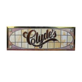 Clyde's Stained Glass Window Panel: A Clyde’s stained glass window panel. This panel features the Clyde’s logo in brown glass over a field of light orange in the shape of an oval. Each corner is made of a purple and orange swirled glass accented with clear glass spheres. The panel is framed in brass with a hinge attached to the top.
