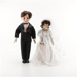 Bride and Groom Porcelain Dolls and Wedding Book: A selection of bride and groom porcelain dolls and a wedding book. This pair features dolls in traditional wedding attire. The Groom is attired in a black tuxedo, a white shirt, dark red bow tie and vest, and a black cumberbund. The Bride is wearing a white wedding dress with lace detail trim to the neck, bodice, cuffs, and hem. This bride doll has a headband with an attached white veil with lace trim and carries a white and pink bouquet. Also included, The Enchanted Dolls’ House Wedding book.