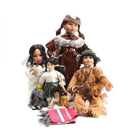 Porcelain Dolls in American Indian Style Outfits and More: A selection of four dolls in American Indian style outfits and more. This group features a porcelain doll wearing a faux suede dress with fringe, beads and a faux fur accent at the shoulders. She wears a beaded necklace with a feather hanging from it and has a woven headband with a single white feather at the front. Her long dark hair is done into pigtails also tied with woven bands. Included with her are an angel doll accented with feathers and beads and with a long dark braid down her back, a doll in a faux suede pant and shirt outfit with beads, braids in her hair and faux fur accents, as well as many more.