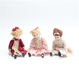 Set of Three Porcelain Dolls: A selection of three dolls in dresses. Each has a porcelain head with inset glass eyes and painted facial features under curly hair, and porcelain hands and lower legs with a body made of cloth. There is a wire armature running throughout so the dolls can be posed, and each is dressed in delicate clothing with lace accents and faux pearl buttons.