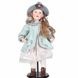 Porcelain Doll with Stand: A porcelain doll with stand. This doll features long brown hair, brown eyes, and rosy cheeks. The doll is dressed in a white dress with a green coat, dark brown shoes, and a grey hat. Included is a stand.