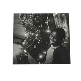 Jill Krementz Black and White Photograph of a Young Girl: A black and white photograph of a young girl by listed American photographer Jill Krementz (b. 1940). Depicted is young girl with an exuberant smile standing in front of a hazy Christmas tree. The work remains unsigned, unmounted and unframed. Inventory numbers to the verso read, “8-112/ 100/ #9-59.”