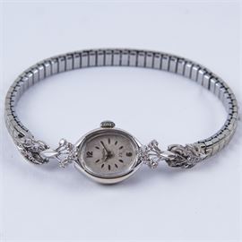 14K White Gold and Diamond Bulova Wristwatch: A 14K white gold and diamond Bulova wristwatch. It features silver tone Arabic numerals and indication bars on a light gray oval face. This rests behind glass in a silver tone housing with pierced, diamond-studded floral lugs. The silver tone expanding band includes floral elements with a single diamond on each end.