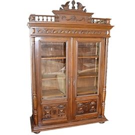 Victorian Style Knockdown China Cabinet: A Victorian style knockdown cabinet. This cabinet features an upper pediment with urn finial above a spindle surround to the top. Below are half fan carvings above two glass paneled doors that open to interior shelving. The cabinet doors are flanked by carved wood columns. The pieces rests on bun feet.