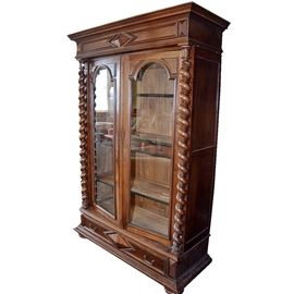 Victorian Style Cabinet: A Victorian style cabinet. This cabinet features a molded top above a diamond shaped carving to the center top. Two cabinet doors with arched glass panels open to three interior wood shelves and three horizontal side by side drawers with dovetail joinery. The cabinet doors are flanked by barley twist columns. Below the cabinet doors is a large drawer with coordinating diamond shaped carving to the center and two bail pulls. The piece rests on bun feet.
