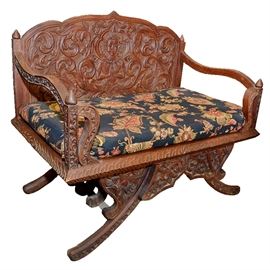 Thai Carved Mahogany Settee: A Thai carved mahogany settee. This piece features an intricately carved back with swirling flourishes and the image of a deity. The carved arms undulate forward meeting carved square supports with carved S-curved accent rails. The seat sides and front are carved, with an upholstered cushion in blue fabric with a pink, yellow, and green floral pattern. The piece features intricately carved front and back skirts and stands on four carved splayed legs.