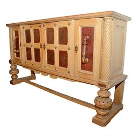 Large French Oak Buffet With Inlaid Tooled Leather Panels: A large French oak buffet with inlaid tooled leather panels. This piece has a light finish. It features two hinged doors to the center with four recessed panels in each. The panels hold burgundy leather inlays with tooled lions and floral motifs highlighted in gold tone. The two doors have gold-toned pyramid shaped nail head accents at each corner and joint. Within are two dovetail jointed drawers above storage shelves. The center doors are flanked by two hinged recessed panel doors with burgundy leather inlays and large gold-toned twist turned vertical handles. Additional storage is within. The front corners feature twisted columns above the turned front legs, which are embellished with small gold-toned pyramid shaped nail head accents. The legs fix to trestle style bases with a beam stretcher. The piece is labeled “Federation Nationale De L’Ameublement/0027” to a door interior. The key is included.