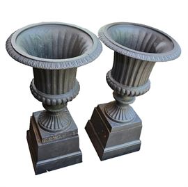 Cast Iron Pedestal Urn Planters: A pair of pedestal urn planters. These pieces are composed a reeded urn shaped pot on a square base. Each is made of cast iron with a black painted finish.