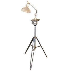Found Object Floor Lamp: A found object floor lamp. This floor lamp is comprised of found objects such as a clear glass shade, a shower head fixture as an arm, and a metal surveying tripod with wooden folding legs. It has one light socket and powers on by a pull chain. A maker’s mark is not present.