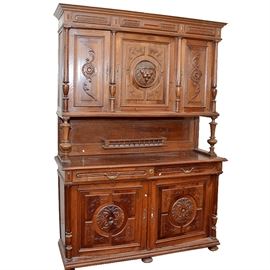 Henri II Style Buffet a Deux Corps: A Henri II style buffet a deux corps. This walnut piece features intricately carved details. It features a molded top with carved detail and raised panel accents above three carved doors set between four turned columns. The center door has a carved protruding lion head, and the outer two doors have carved sunflowers. Storage shelves are within. The upper unit stands on two turned posts to the front and an incised back panel, with a gallery shelf to the center with spindles and a top rail. The lower unit has two locking side-by-side dovetail jointed drawers with brass-toned lion head bail handles. Below are two raised panel doors with carved floral medallions. The doors open to reveal additional storage shelves. The doors are flanked by turned columns, fixed to a molded base. The piece stands on bun feet. The keys are included.