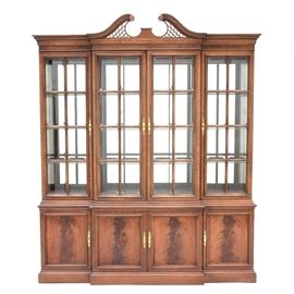 Mahogany Finish Breakfront China Cabinet: A mahogany finish breakfront china cabinet. Comprised of two pieces; the lighted top section features a divided arch pediment over four doors with beveled glass panes and adjustable glass shelving. The base has four cabinet doors with flame mahogany veneers, opening to shelved interior storage, on a stepped plinth base.