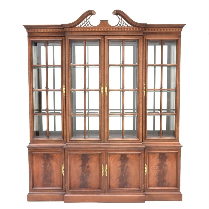 Mahogany Finish Breakfront China Cabinet: A mahogany finish breakfront china cabinet. Comprised of two pieces; the lighted top section features a divided arch pediment over four doors with beveled glass panes and adjustable glass shelving. The base has four cabinet doors with flame mahogany veneers, opening to shelved interior storage, on a stepped plinth base.