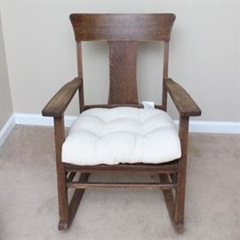 Antique Oak Rocking Chair Circa 1900s: An antique oak rocking chair circa 1900s. The chair is oak with a walnut finish and displays a slat back and wide crest rail. Other features include straight arms and open sides, a wood seat and a button tufted loose seat cushion that sits on straight wood block legs on runners.