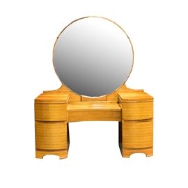 Vintage Blonde Tone Wood Vanity Table With Round Mirror: A vintage blonde tone wood vanity table with a round mirror. The table features: a large round mirror over the center of the table, which has a slightly inward curved front and straight apron above the knee hole, with sets of two bowed front stacked drawers elevated above the center to either side. Each of the drawers has a recess underneath it to reach in and pull it open.