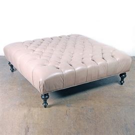 Tufted Leather Ottoman: A large button-tufted leather ottoman. This ottoman features a square shaped padded top upholstered in a tan colored leather, which is attached to four turned legs in a dark stain. No maker’s marks are present.