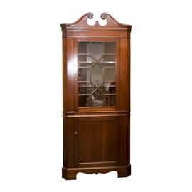 Traditional Style Corner Cabinet: A traditional style corner cabinet. The dark stained wood cabinet features broken scroll pediment at the front of the triangular top, a glass front door embellished with wood accents arranged in a diamond and cruciform pattern, a wood door below the glass door, a scroll cut apron, and bracket feet. The cabinet has two fixed shelves behind the glass door and one fixed shelf behind the wood door. Each door has a brass tone key plate, no key is included.