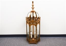 Over-sized Copper-tone Footed Lantern: An oversized copper-toned footed lantern. This electric lantern features a domed top with long finial, atop a floral scrolled base with glass sides. There are six candle lights and one two-prong cord. Companion item 17BAL076-012