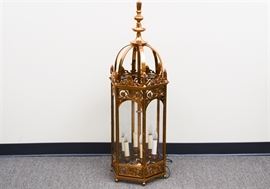 Over-sized Coppertone Footed Lantern: An oversized copper-toned footed lantern. This electric lantern features a domed top with long finial, atop a floral scrolled base with glass sides. There are six candle lights and one two-prong cord. Companion item 17BAL076-004.