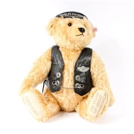 Steiff Teddy Bear Harley-Davidson 100th Anniversary Edition: A Steiff teddy bear commemorating Harley-Davidson’s 100th anniversary. The blonde teddy bear has brown glass or plastic eyes, with a black stitched nose and mouth. The bear wears a black “Harley-Davidson” cap and faux leather vest with silver buttons, chains and trim. The bear’s feet are embroidered with “1903-2003” to the right sole, and with “100 Years of Great Motorcycles” to the left sole. The bear’s left ear has a Steiff button and tag, and there is a paper tag attached to the left wrist. A fabric label attached to the wrist is marked “666476 – L. Ed. of 5,000 pcs. – 02240”. Also included is a certificate of authenticity from Harley-Davidson Motor Company and Margarete Steiff GmbH", certificate number “002240”.