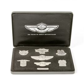 Nickel Silver Harley-Davidson Medallions: A collection of nickel-silver Harley Davidson medallions. Featured here is a collection of medallions commemorating the 50th, 75th, 80th, 85th, 90th, and 100th anniversary of Harley-Davidson. All of the insignias come displayed in a black felt lined tin box. The felt has a plaque attached with the number 1043/2003 and there is a certificate of authenticity noting that the medallions are made of nickel silver. Beneath the medallion tray, in the bottom of the box are a pair of black cloth gloves. The exterior of the tin box is finished in black and marked “100 years of great motorcycles, 2003” with the Harley-Davidson logo.
