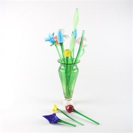 Glass Flower Bouquet in Glass Vase: A bouquet of art glass flowers in a glass vase. The bouquet includes flowers in colors of blue, yellow, cobalt, ivory, and red. They are presented in a green tint amphora vase with striated brown accents and clear pedestal base.