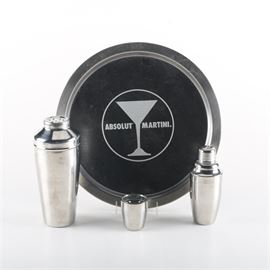 Stainless Steel Bar Set: A stainless steel bar set. Included are two shakers in small and large sizes, one shot glass, and a round tray with martini glass and “Absolut Martini” to the front. The attached sticker to the underside of the tray reads “Vonpok Made in Taiwan”.