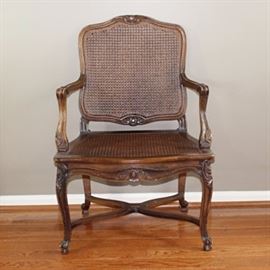 Louis XV Style Cane Armchair: A Louis XV style cane armchair. The chair features a slightly arched square back with cane support, curved armrests, a cane seat and cabriole legs on whorl feet with an X stretcher. There are carved rosettes and circular reliefs decorating the arms, legs, and apron.