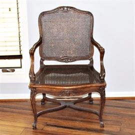 Louis XV Style Cane Armchair: A Louis XV Style cane armchair. It features a slightly arched square back with cane support, curved armrests, a cane seat and cabriole legs on whorl feet with an X stretcher. There are carved rosettes and circular reliefs decorating the arms, legs and apron.