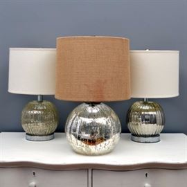 Mercury Glass Table Lamps: A group of three mercury glass table lamps. The matched pair features fluted bulbous bodies, chrome bases, and white fabric drum shades. The larger electric light features a bulbous mercury glass body and includes a burlap fabric shade.