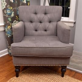 Upholstered Club Chair: An upholstered club chair. This chair features a square back with button tufting over a removable square seat with button tufting flanked by high arms with silver toned nailhead trim to the exterior. The chair rises on turned legs terminating in arrow feet. The chair is upholstered in a grey toned fabric. This item matches 17IND083-005.