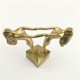 Art Deco Style Brass Statue of Women: An Art Deco style brass statue of women. This freestanding statue depicts two slender, matching women interlocked in a mirrored form. They arch their backs while holding onto each other’s wrists while standing on a wedge base featuring gouged, linear lines to the edges. To the underside is an affixed felt pad for support. Unmarked.