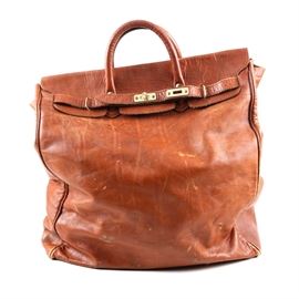 "Vaca Lisa" Genuine Leather Tote Bag: A large brown leather tote bag. The features an orangish brown genuine leather tote with two handles, gold tone hardware and a buckle strap across the front. The bottom has five stud feet and a suede interior with a label that reads “Genuine Leather, Made in Argentina, Vaca Lisa”.