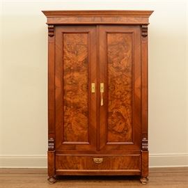 Empire Style Burled Armoire: An Empire style burled armoire. This piece features a mahogany finish with burl and inlay details. To the top is crown molding, above a pair of recessed panel doors flanked by fluted columnar trim. The doors feature rectangular brass-toned escutcheons and a skeleton key with a gold hued rope and tassel. Below the doors is a single full-length drawer with a recessed panel front and a brass-toned bail handle. The armoire stands on bun feet.
