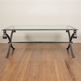 Glass Top Coffee Table: A glass top coffee table. This table features a rectangular top with a framed panel of clear glass over a metal X motif trestle frame. The sides include ball and scrolling and the center trestle support includes twisting and hammered metal design elements.