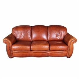 Leather Sofa by Distinctions Leather: A leather sofa by Distinctions Leather. This sofa features a scalloped cushion back and rolling arms flanking removable cushion seats with scalloped front seat support rail and nailhead trim. The sofa rests on wooden bun feet, is upholstered in a raven wood chestnut tone leather and is marked “Distinctions Leather".