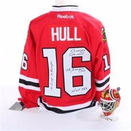 Signed Bobby Hull Blackhawks Jersey and Mug: A signed Bobby Hull Blackhawks jersey and mug. This selection includes a jersey and helmet-shaped mug that each showcases the Blackhawks decal and the signature of Bobby Hull. The mug is numbered A 0781 59/299. These items are labeled by the manufacturers.