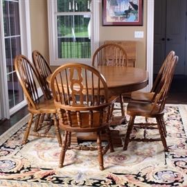 Oak Dining Table and Six Windsor Style Chairs by Keller: An oak dining table with six Windsor style chairs by Keller. This features one oak table, round or oval depending on use of the single leaf; it sits on a pedestal base with two turned columns and is marked "Keller’ with a quality tag to the underside. Also includes are six English Windsor style chairs; two are sack-back armchairs with a spindle and pierced splat back while the other four are bow-back with similar splats. All sit on turned legs with an H-stretcher between them. They are marked to the underside.