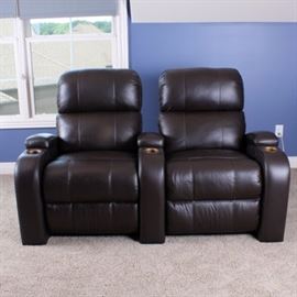 Pair of Reclining Theater Seats: A pair of reclining theater seats. Each seat features a brown faux leather upholstery and an overstuffed pillow design with an electronic reclining back and lifting leg-rest. One of the seats includes two-panel armrest sides and the other includes one with gold tone cup holder inserts and pillow rests.