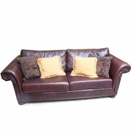 Leather Sofa with Brass Tack Detail: A leather sofa with brass tack detail. Featured is a brown leather sofa with carved wood armrests and apron, rolled arms, brass rivet details, and bun feet. Includes four accent pillows.