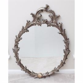 Ornate Wall Mirror: A wall mirror in an elaborate decorative style resin frame. This oval mirror boasts a silver-tone accented frame with sharp, curvaceous embellishments throughout. Mirror hangs by a wire to verso.