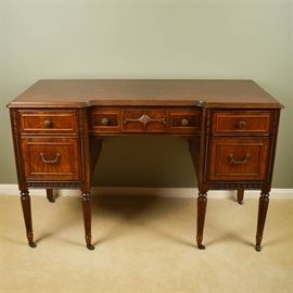 Vintage Federal Style Walnut Writing Desk: A vintage Federal style writing desk. The desk is constructed of wood and walnut veneer with burl details. The desk offers a single upper center drawer flanked by two drawers to each side. Each has ornate knobs and pulls with paneled drawer fronts and dovetail joinery. The desk has ornate applied trim throughout and rises on reeded tapered legs that terminate into small metal casters. The desk retains a partial maker’s label to the back of the piece. Matches chest of drawers listed separately as item 17CIN384-040.