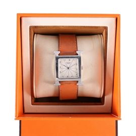 Stainless Steel Hermes Wristwatch: A stainless steel Hermes wristwatch. This features a silver tone square bezel in the shape of an “H” with a white square dial that has a crossing line design throughout with black Arabic numeral hour indicators and slender silver hands that leads to a brown leather band. A bright orange “Hermes” case is also included.