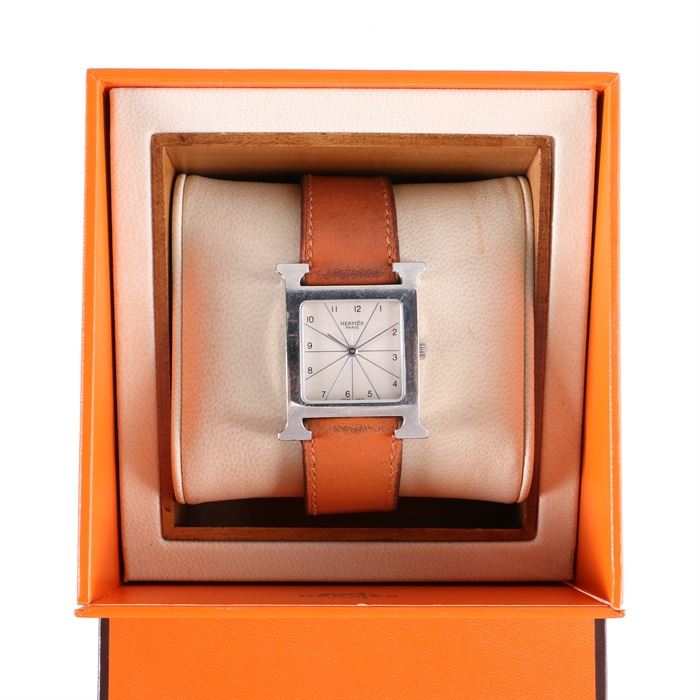 Stainless Steel Hermes Wristwatch: A stainless steel Hermes wristwatch. This features a silver tone square bezel in the shape of an “H” with a white square dial that has a crossing line design throughout with black Arabic numeral hour indicators and slender silver hands that leads to a brown leather band. A bright orange “Hermes” case is also included.