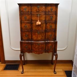 Early 20th Century Burl-Front Highboy: An early 20th century burl-front highboy. This piece features a molded cornice over a face with burled veneer and four graduated, hand-dovetailed drawers. Each wave-front drawer features ring pulls with a circular backplate and escutcheon to the center; keys are included, one with a large tassel attached. The base features one similar drawer, leading to a serpentine apron and cabriole style legs with scroll detailing and pad feet.